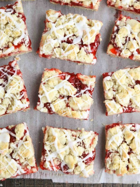 Squares of glazed cherry pie bars on parchment paper and a cooling rack.