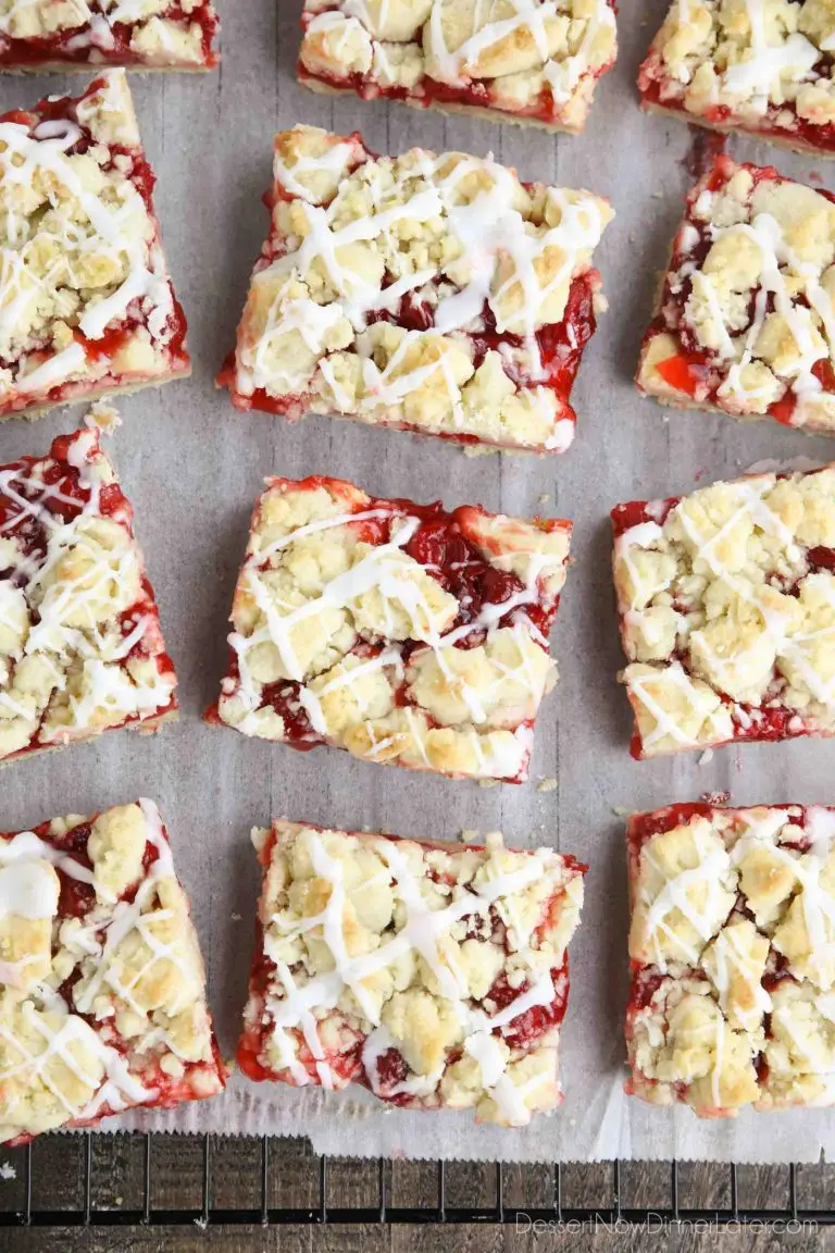 Squares of glazed cherry pie bars on parchment paper and a cooling rack.