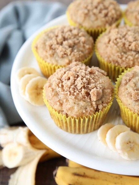 White cake plate with banana muffins in yellow wrappers with crumb streusel on top and slices of banana on the sides.