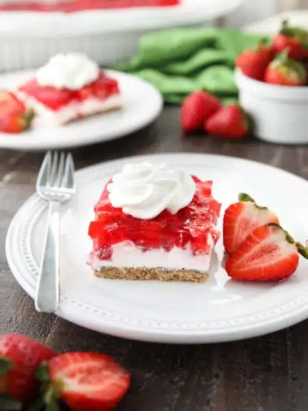 A square of strawberry delight on a plate. (A layered dessert with a graham cracker crust, no bake cheesecake filling, and a layer of fresh strawberries and jello with a swirl of whipped cream on top.)