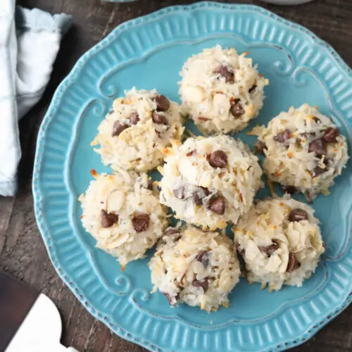 Top view of Almond Joy Cookies (chocolate almond macaroons) on a plate with bowls of chocolate chips and sliced almonds on the side.