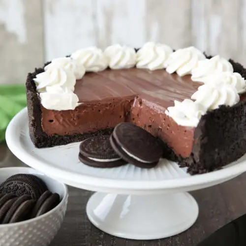 No-bake chocolate cheesecake and Oreos on a cake plate with whipped cream piped on top.