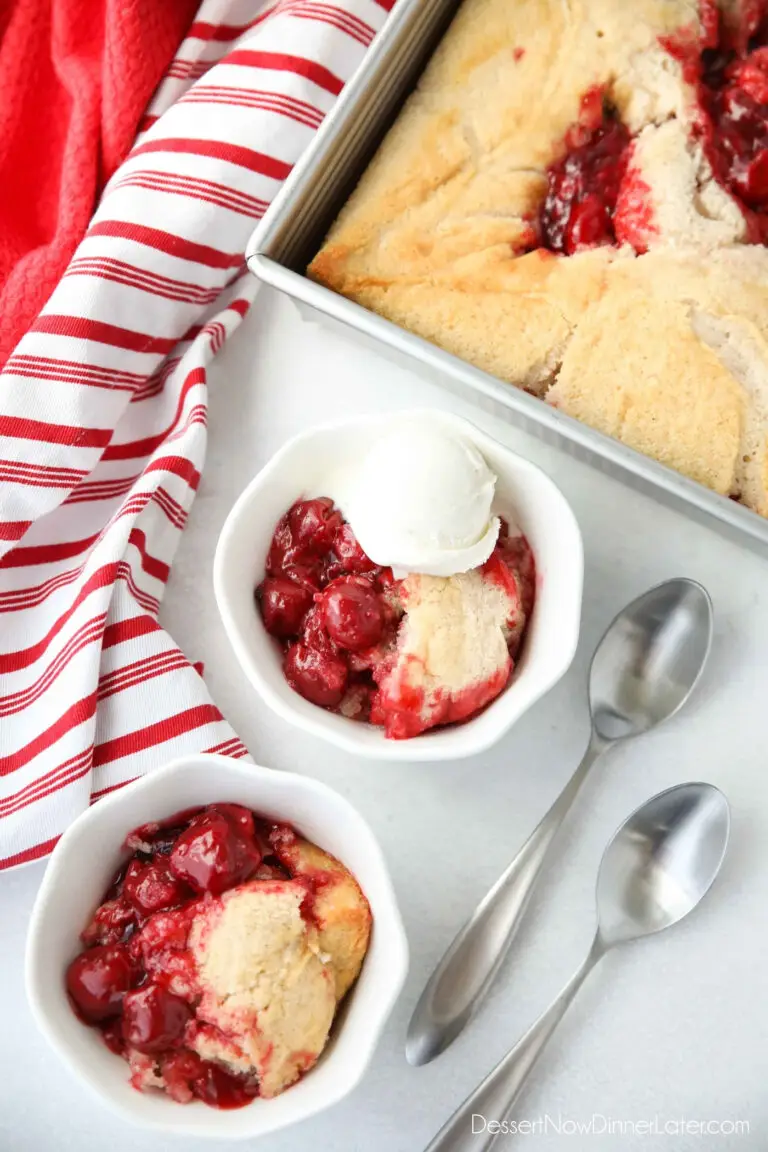 Two bowls of easy cherry cobbler with two spoons on the side. One serving of cobbler has ice cream on top, and is sitting next to the pan it was baked in.