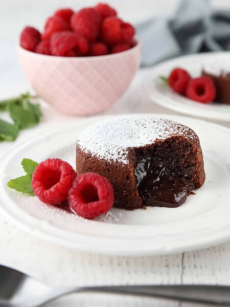 Molten chocolate lava cake on plate dusted with powdered sugar and a side of raspberries and mint, with gooey center melting onto the plate.