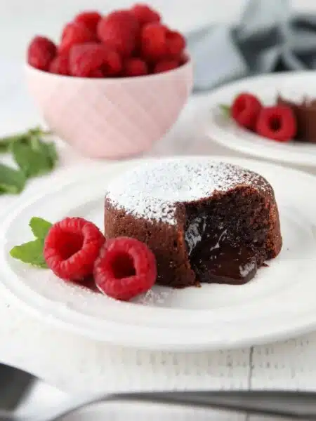 Molten chocolate lava cake on plate dusted with powdered sugar and a side of raspberries and mint, with gooey center melting onto the plate.