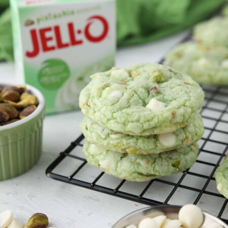 Three pistachio cookies with white chocolate chips stacked on top of each other. Box of pistachio pudding in the background.