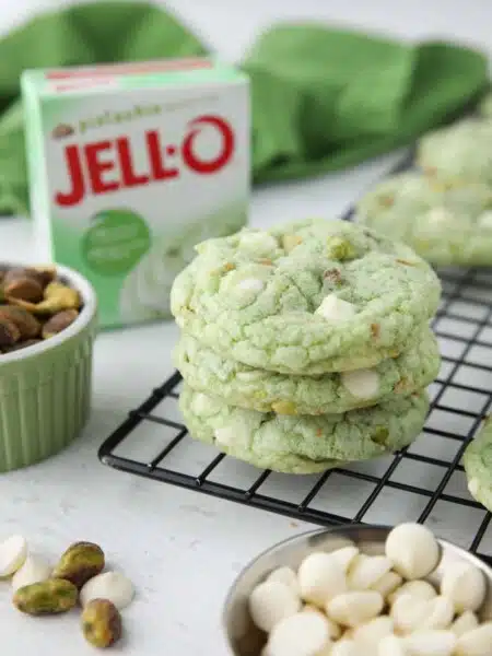 Three pistachio cookies with white chocolate chips stacked on top of each other. Box of pistachio pudding in the background.