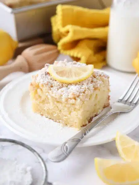 Thick slice of lemon coffee cake on a plate dusted with powdered sugar and a slice of lemon on top.