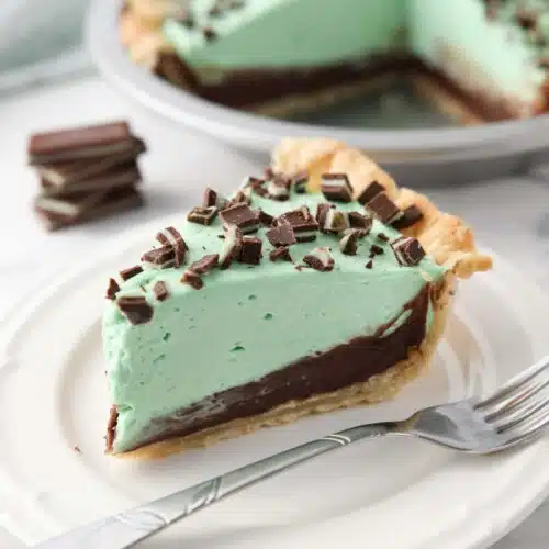 Slice of chocolate mint pie on a plate with layers of pastry crust, chocolate ganache, mint no bake cheesecake, and chopped Andes mints.