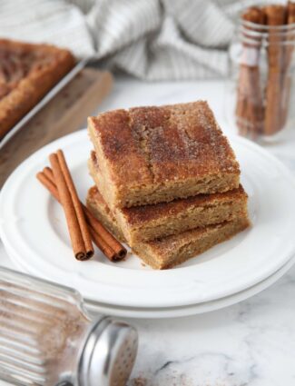 Stack of cinnamon bars on a plate with extra cinnamon-sugar on top and cinnamon sticks on the side.