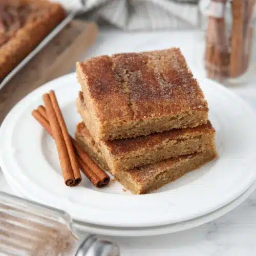 Stack of cinnamon bars on a plate with extra cinnamon-sugar on top and cinnamon sticks on the side.