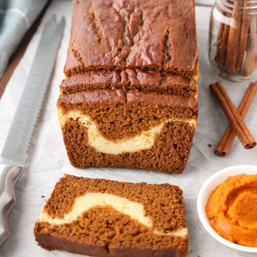 A loaf of pumpkin cream cheese bread with a few slices cut, showing a swirl of cream cheese in the center.