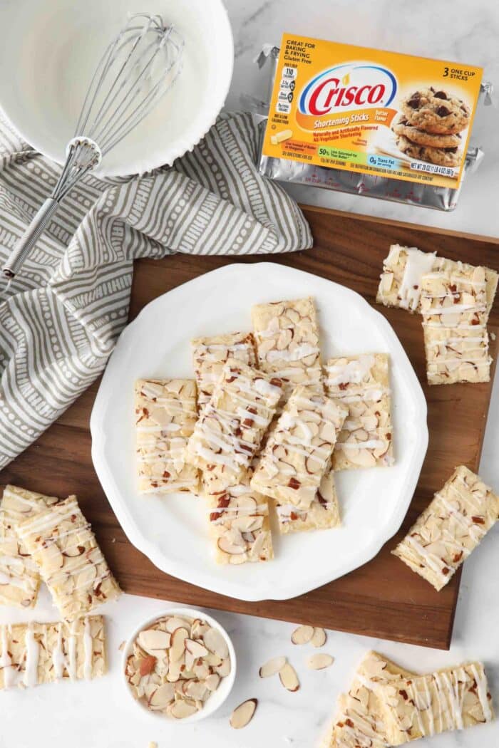 Top view of almond bars on a plate with Crisco shortening, sliced almonds, and a bowl of glaze on the side.