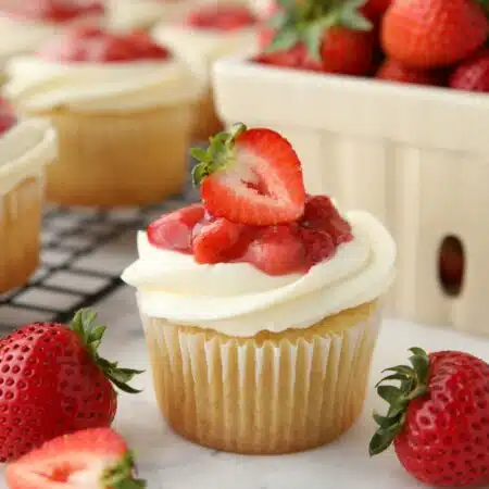 Close up of a Strawberry Shortcake Cupcake with vanilla cake, whipped cream cheese frosting, and strawberry sauce on top with a strawberry garnish.