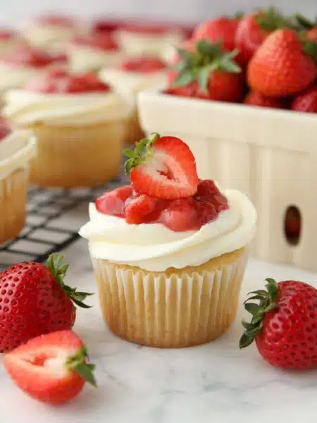 Close up of a Strawberry Shortcake Cupcake with vanilla cake, whipped cream cheese frosting, and strawberry sauce on top with a strawberry garnish.