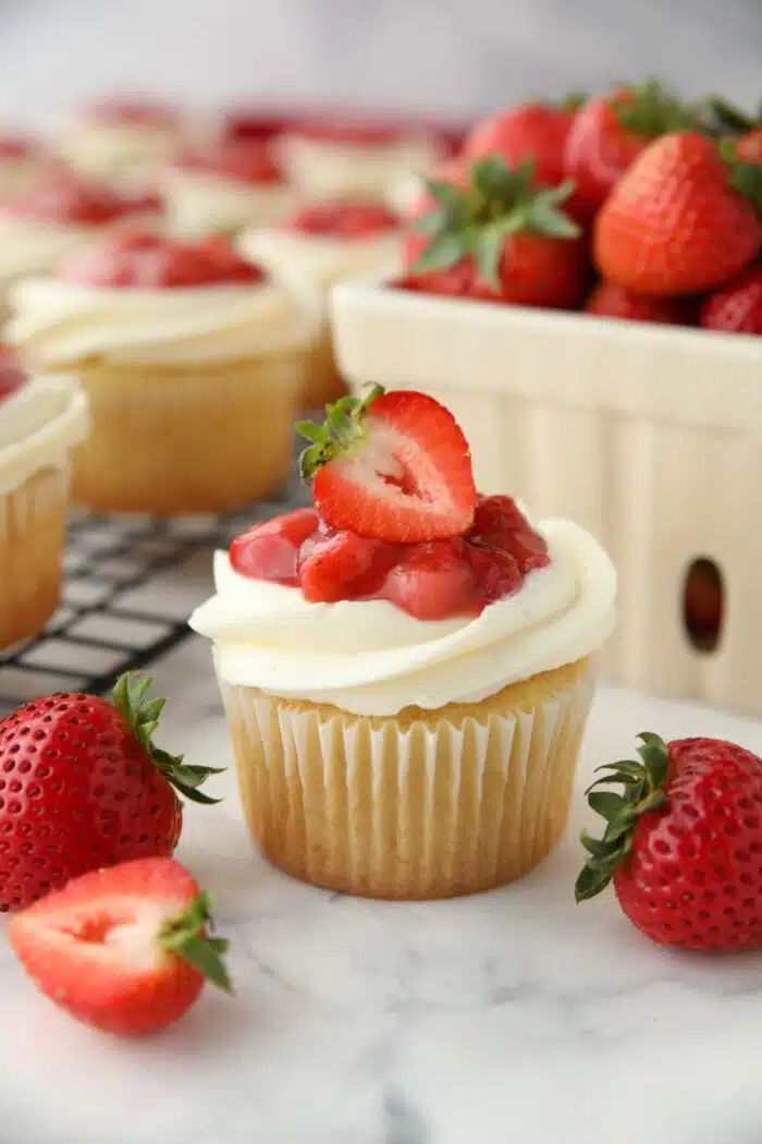 Close up of a Strawberry Shortcake Cupcake with vanilla cake, whipped cream cheese frosting, and strawberry sauce on top with a strawberry garnish.