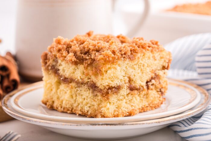 A slice of cinnamon crumb cake on a plate and a cup of coffee in the background.